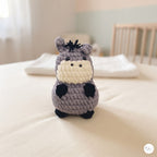 A small crocheted donkey toy sits centered on a white blanket in a softly blurred nursery setting, with a crib and pale walls visible in the background.