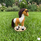 A crocheted toy horse with brown and white coloring stands on a small round wooden base in a grassy outdoor garden setting.