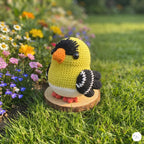 A crocheted bird toy in yellow, black, white, and orange is displayed on a small wooden slice stand in a grassy garden with colorful flowers in the background.
