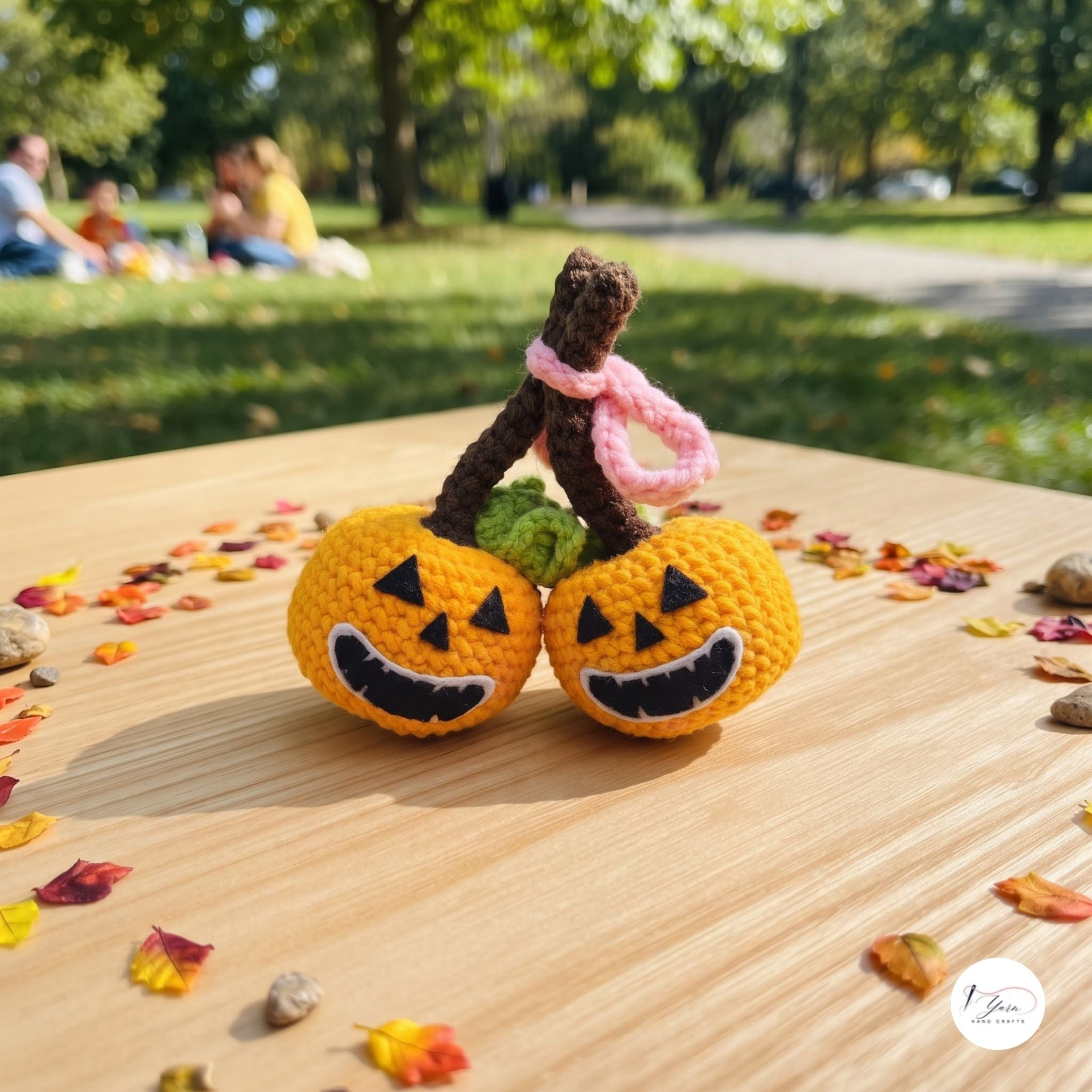 Two small crocheted jack-o'-lantern ornaments with carved black facial features sit on a wooden table outdoors, with a pink loop handle and green stems. A blurred park scene with people is in the background, and scattered autumn leaves surround the table.