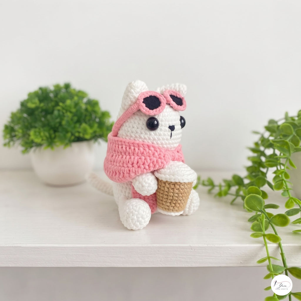 A crocheted white cat plush toy wearing pink sunglasses and a pink scarf, holding a cupcake, displayed on a white shelf with green plants in the background.