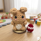 A crocheted plush toy resembling a small brown bunny or bear sits on a wooden surface in a softly lit room. The toy has large black eyes, pink stitched cheeks, a white patch on its front, and two small ears on top.