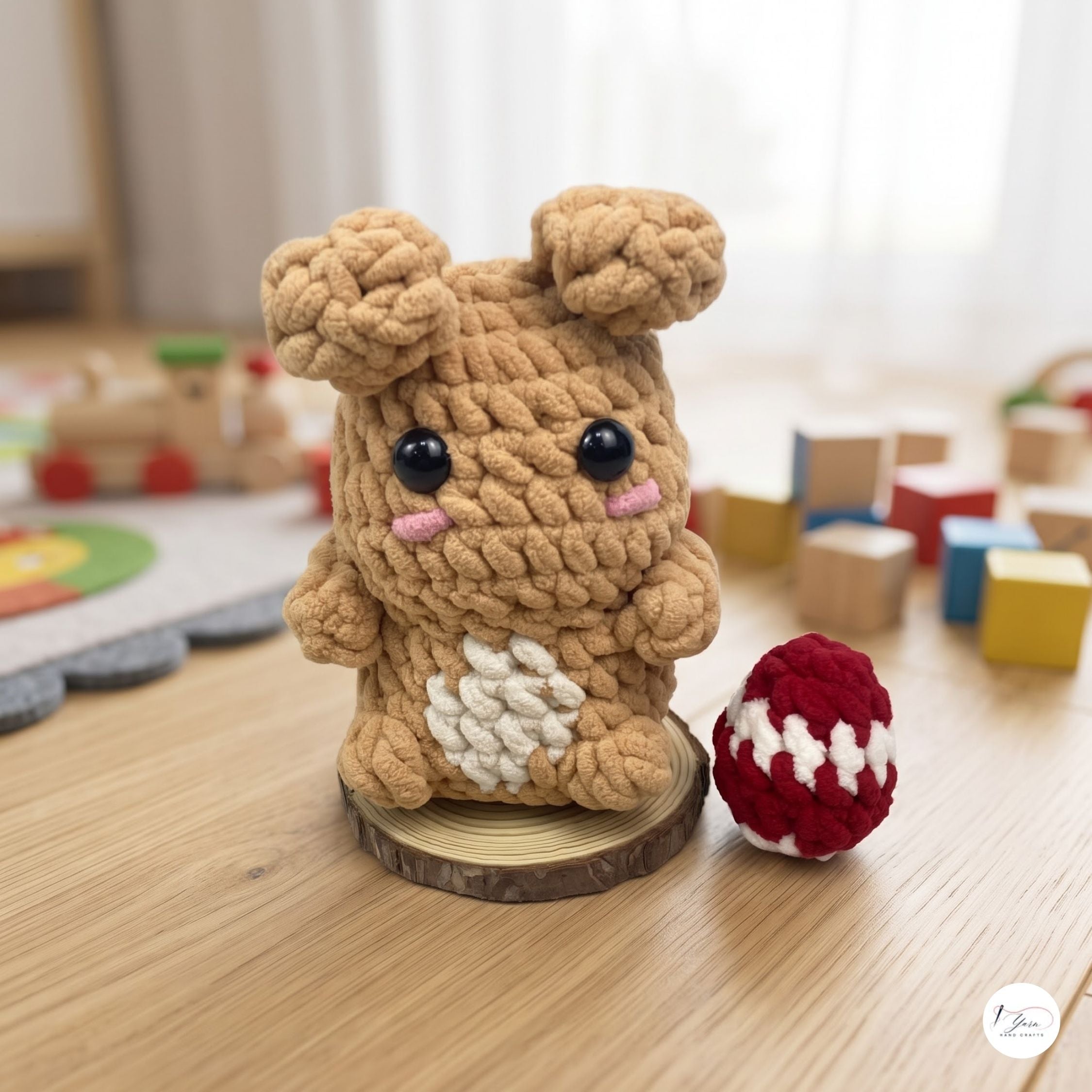 A crocheted plush toy resembling a small brown bunny or bear sits on a wooden surface in a softly lit room. The toy has large black eyes, pink stitched cheeks, a white patch on its front, and two small ears on top.
