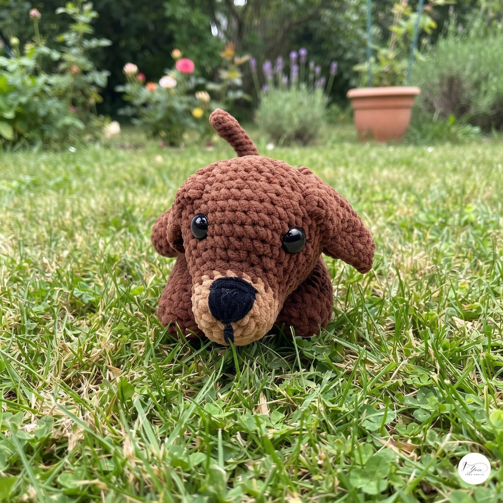 A brown crocheted plush dog toy with black eyes and a beige snout, sitting on a green grassy lawn with a garden background.