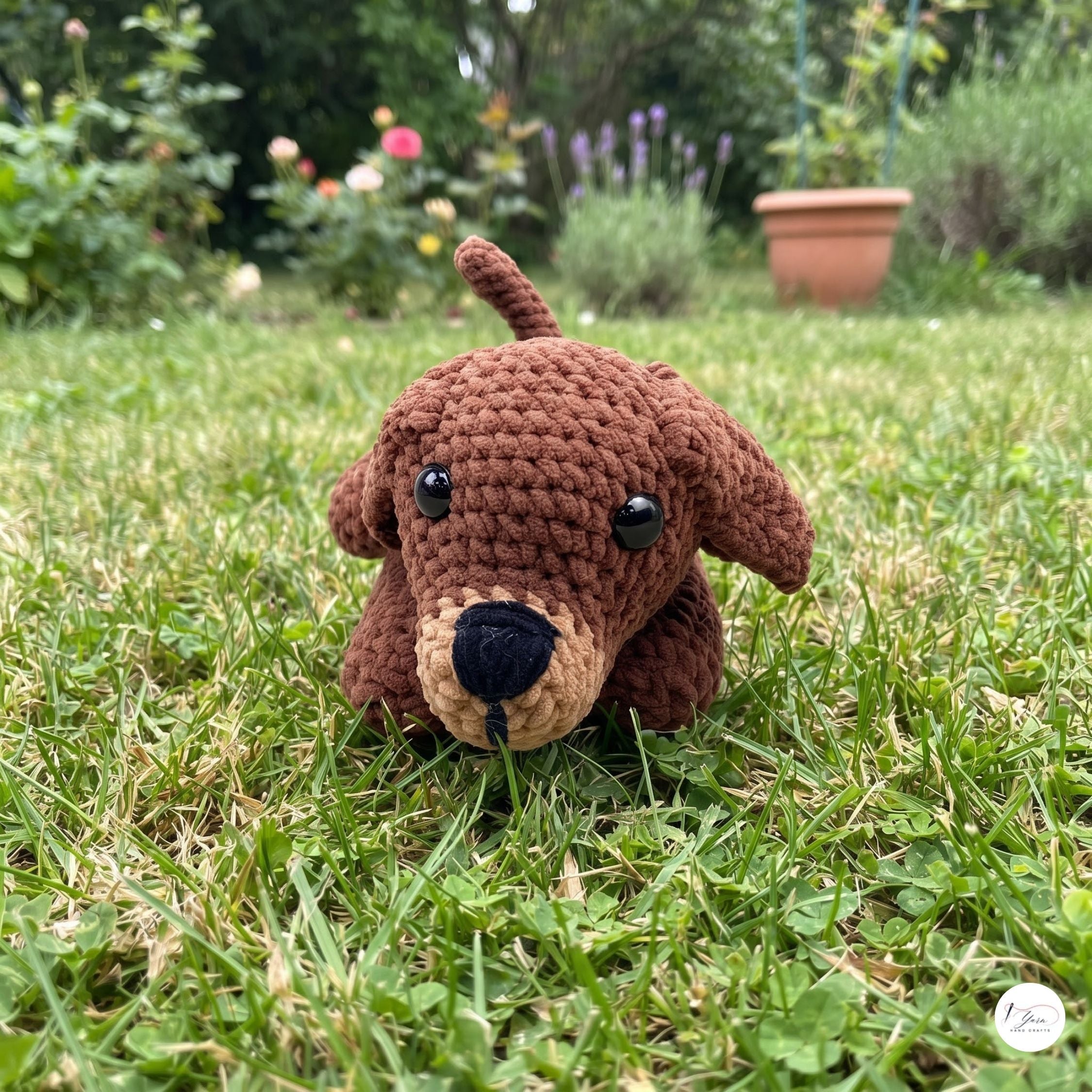 A brown crocheted plush dog toy with black eyes and a beige snout, sitting on a green grassy lawn with a garden background.