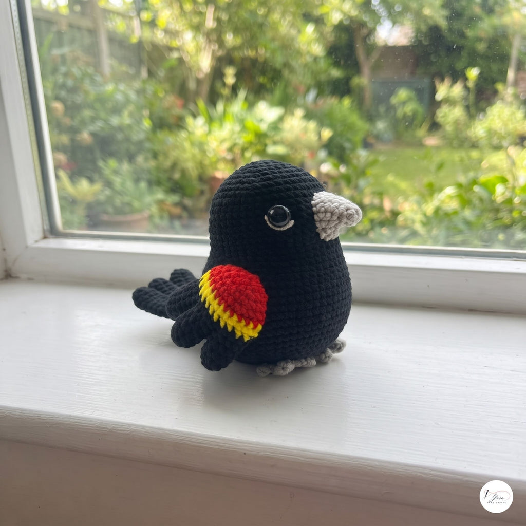 A crocheted bird toy in black with a red and yellow wing patch and gray beak/feet, sitting on a white windowsill with a garden view in the background.