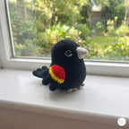 A crocheted bird toy in black with a red and yellow wing patch and gray beak/feet, sitting on a white windowsill with a garden view in the background.