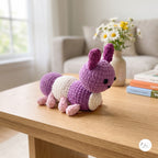 A crocheted plush caterpillar toy in purple, cream, and pink sits on a light wood table in a cozy living room setting with a sofa, flowers, and books in the background.