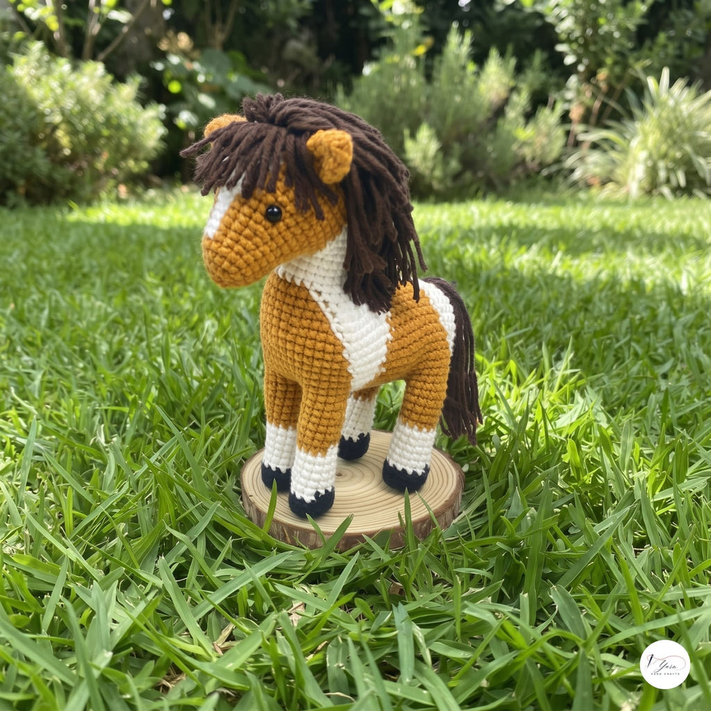 A crocheted plush horse toy with a brown and white pattern, standing on a small round wooden base in a grassy outdoor garden setting.