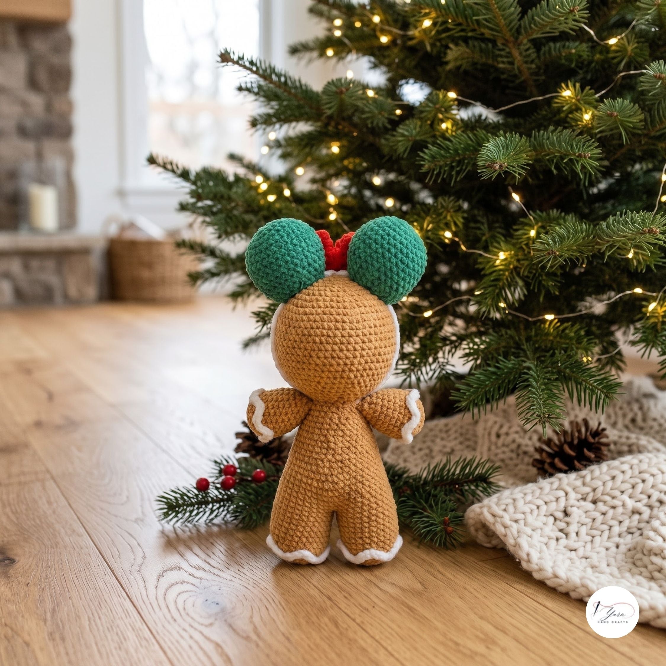 A crocheted Christmas gingerbread doll viewed from behind, standing on a wooden floor beside a decorated Christmas tree with warm string lights. The doll has a tan body, green ears, a small red bow, and white trim.