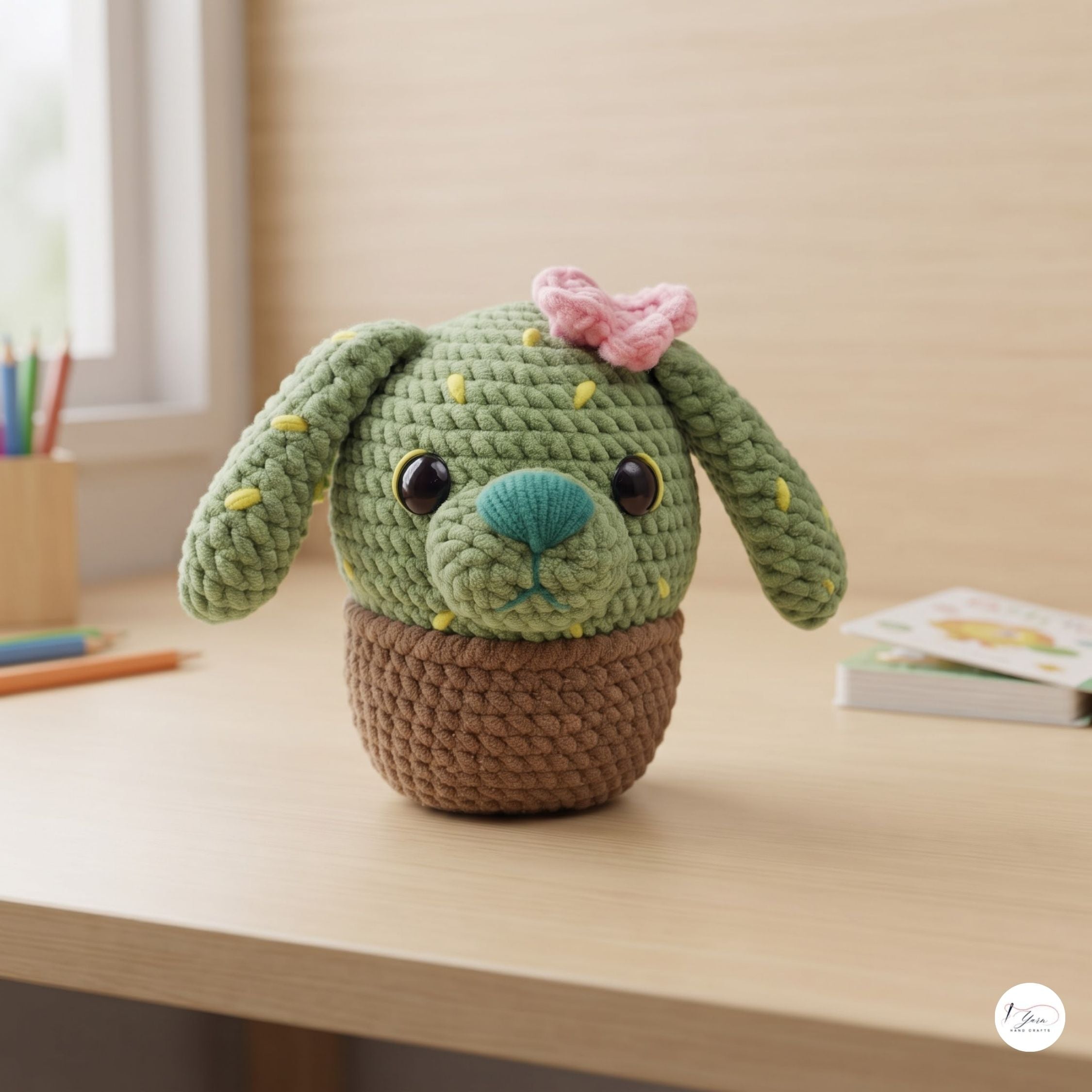 A crocheted plush dog toy with long floppy ears, a pink bow, and a teal nose sits in a brown basket on a light wood desk. The background shows a softly blurred indoor setting with a pencil holder and a small book.
