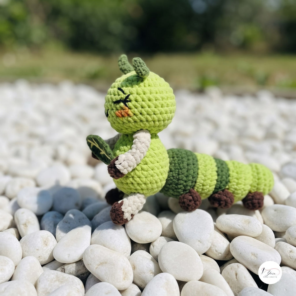 A crocheted caterpillar toy in shades of green, brown, and cream, posed on a bed of smooth white pebbles outdoors with a blurred natural background.