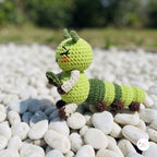 A crocheted caterpillar toy in shades of green, brown, and cream, posed on a bed of smooth white pebbles outdoors with a blurred natural background.