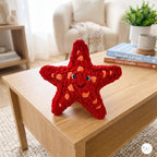 A red crocheted starfish plush toy with orange spots and a smiling face, displayed on a light wood coffee table in a cozy living room setting.