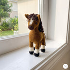 A crocheted horse toy standing on a white windowsill, with a real outdoor garden visible through the window behind it.