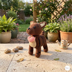 A crocheted stuffed dog toy stands on a stone patio in a garden setting, holding a pink heart-shaped treat. The background features potted plants, a wooden trellis, and a small ceramic teapot.