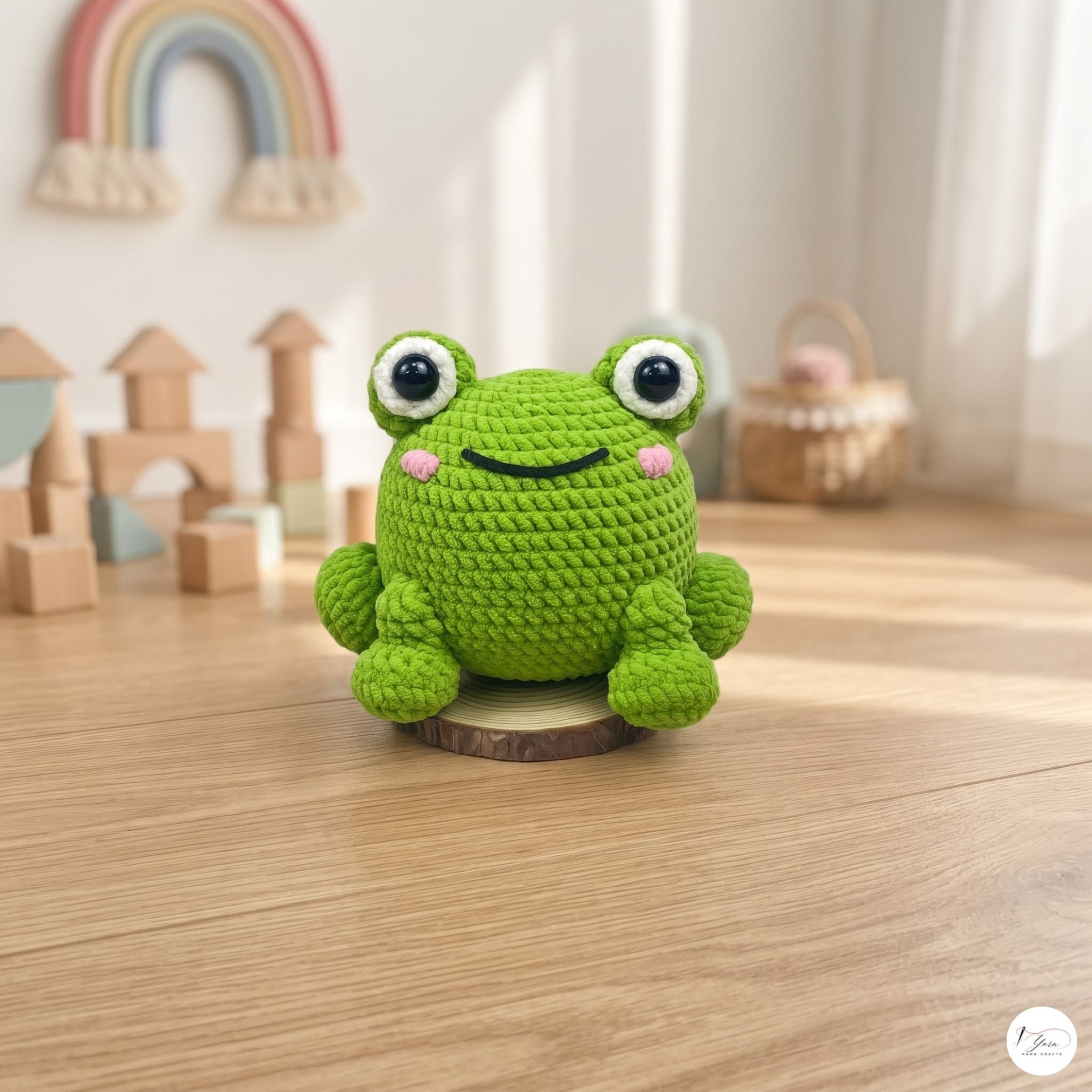 A green crocheted frog toy sits on a small round wooden base on a light wood floor, with a softly blurred children's room background including toy blocks, a rainbow decoration, and a basket.