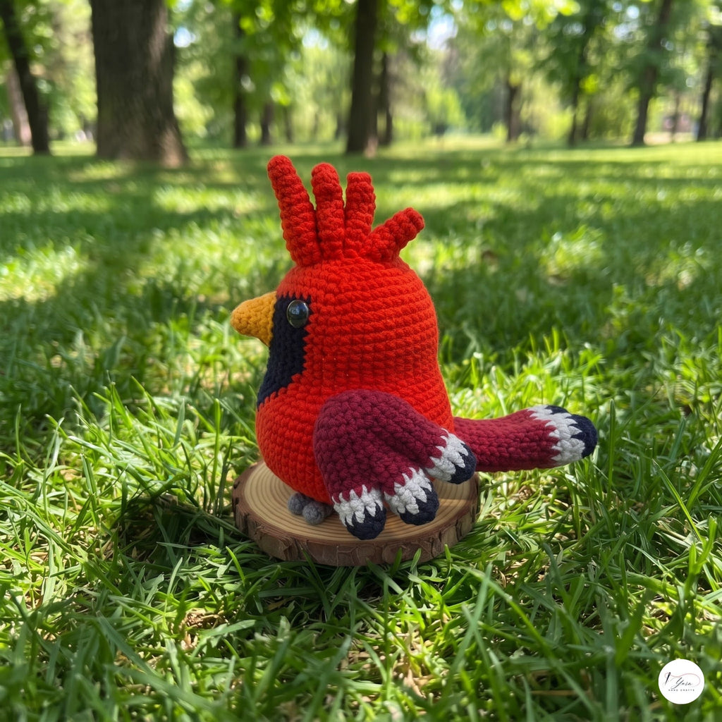 A crocheted red cardinal bird toy sitting on a small round wooden slice in a grassy park with trees in the background.