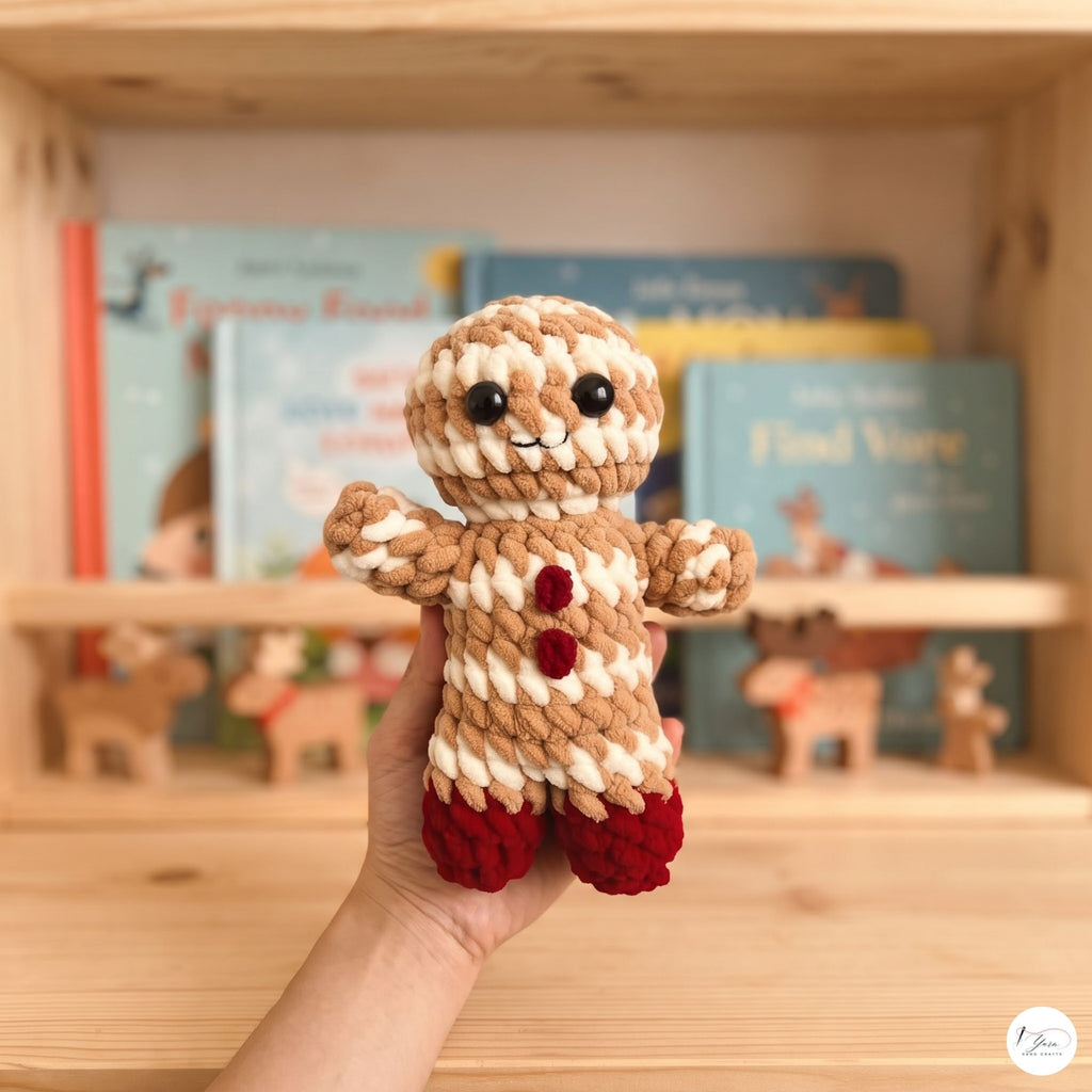 A hand holds a small crocheted gingerbread doll with beige and white striped yarn, red yarn buttons, and red yarn feet in front of a wooden bookshelf with children's books.