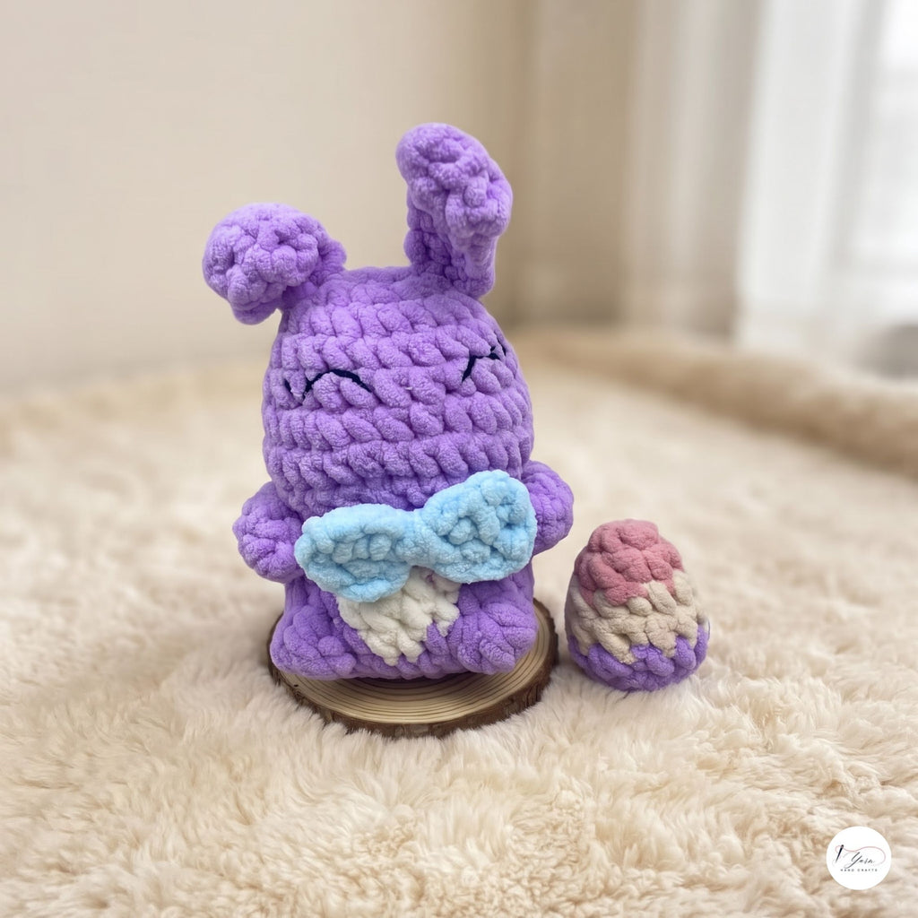 A crocheted purple bunny plush toy with light blue and white wings, sitting on a small wooden coaster next to a matching small pouch, set on a cream-colored faux fur surface.