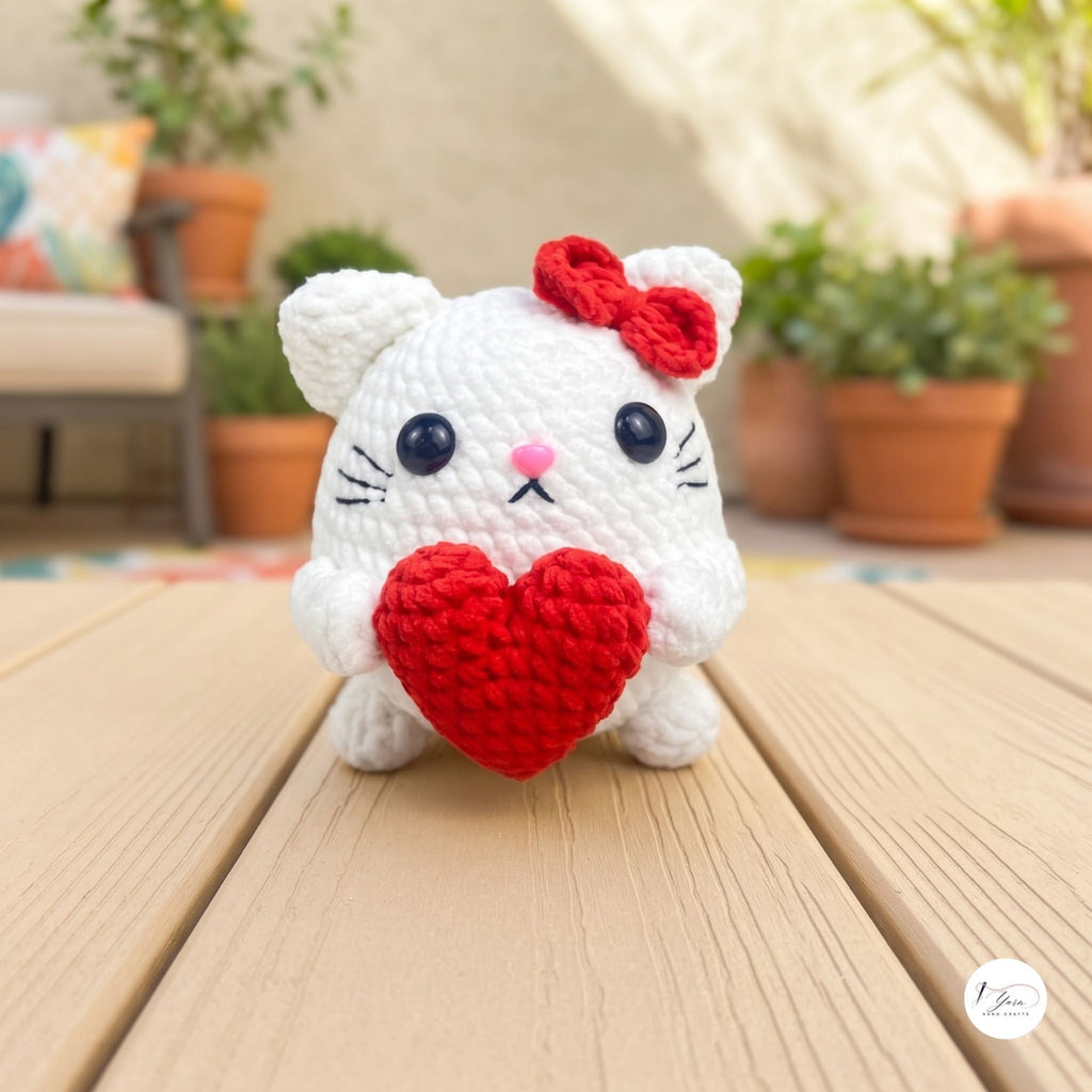 A white crocheted cat toy holding a red heart, with a red bow on its head, set on a wooden deck with a blurred outdoor patio background.