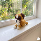 A crocheted dog plush toy in tan, brown, and white sits on a white windowsill with a blurred garden view in the background.