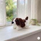 A small crocheted dog toy with brown and white yarn sits on a white windowsill. The toy has long floppy ears, a black nose, and a black bead eye.