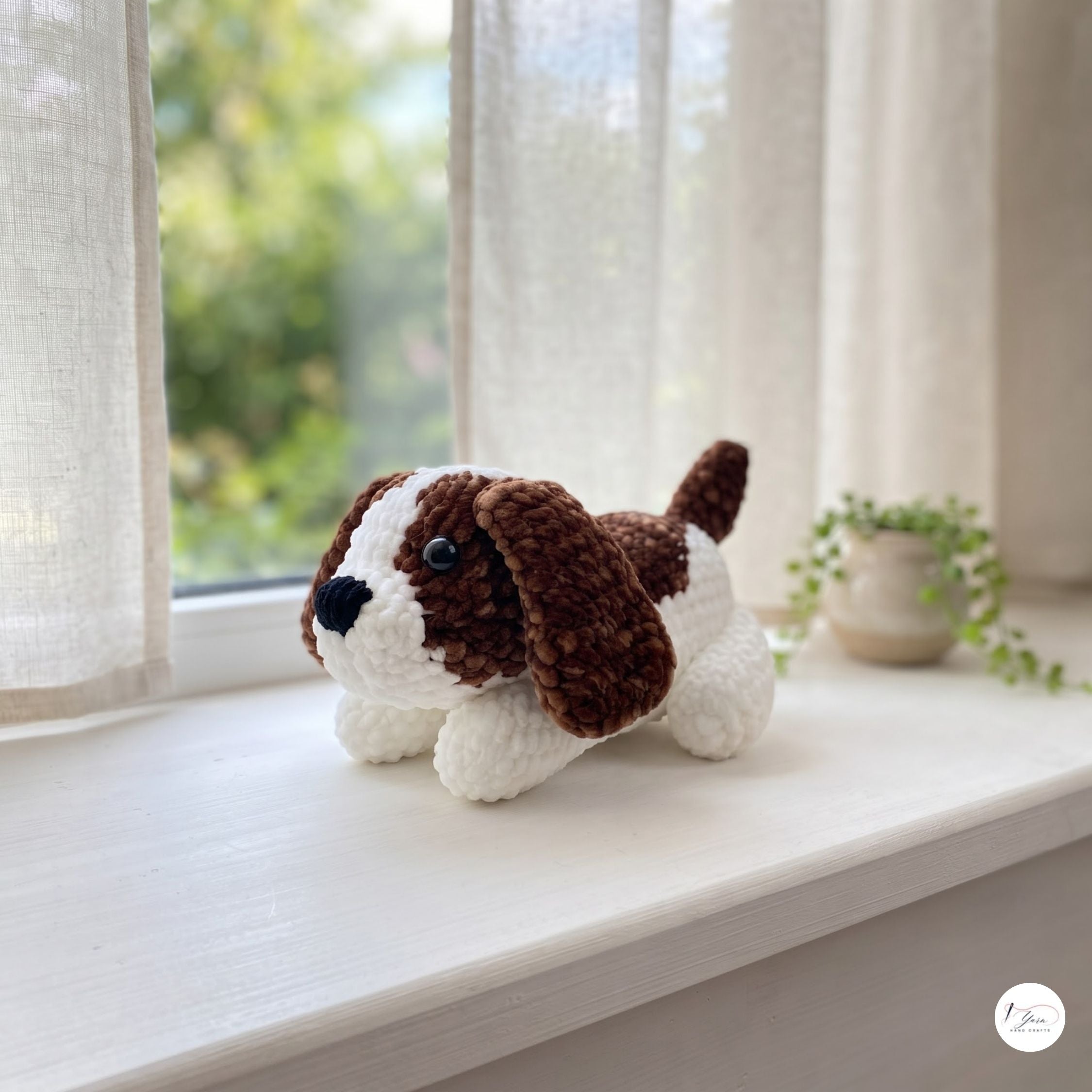 A small crocheted dog toy with brown and white yarn sits on a white windowsill. The toy has long floppy ears, a black nose, and a black bead eye.