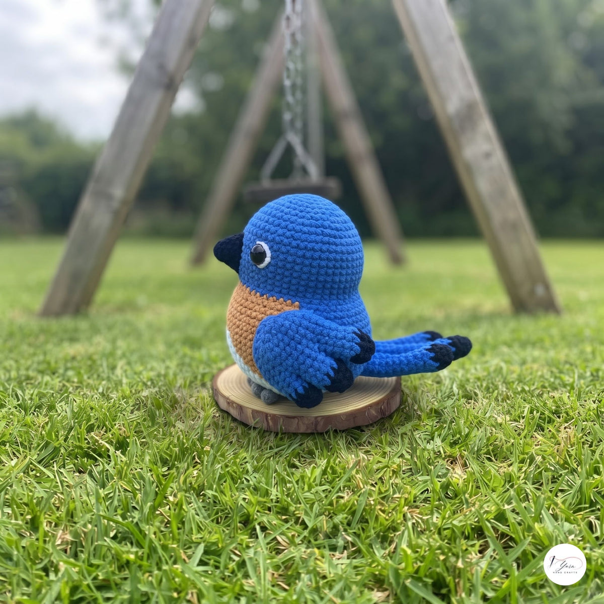 A crocheted bluebird ornament sits on a small round wooden base on a grassy lawn, with a blurred outdoor swing set in the background.