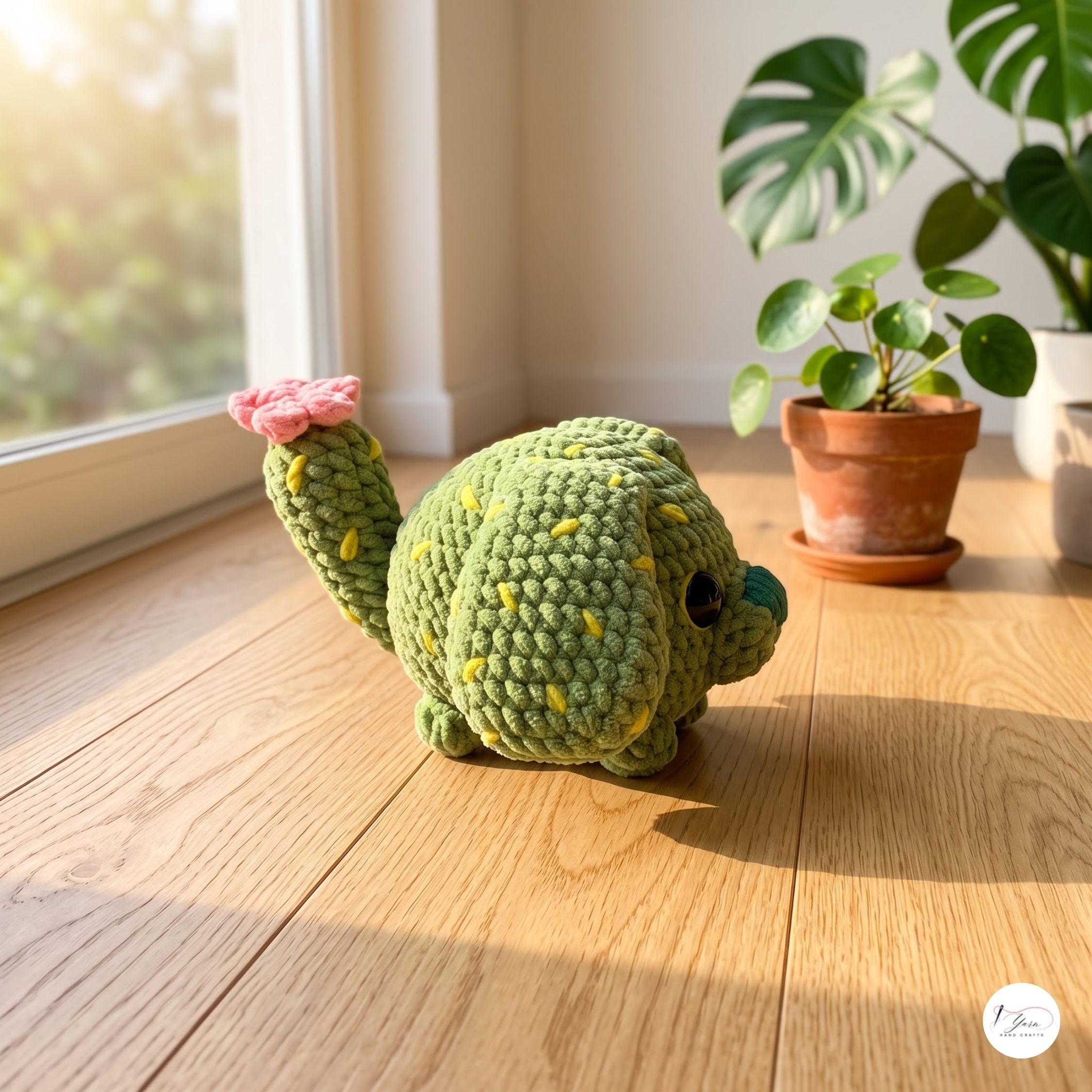 A green crocheted cactus plush toy with yellow spines and a pink flower on its arm, placed on a wooden floor near a window with potted plants in the background.