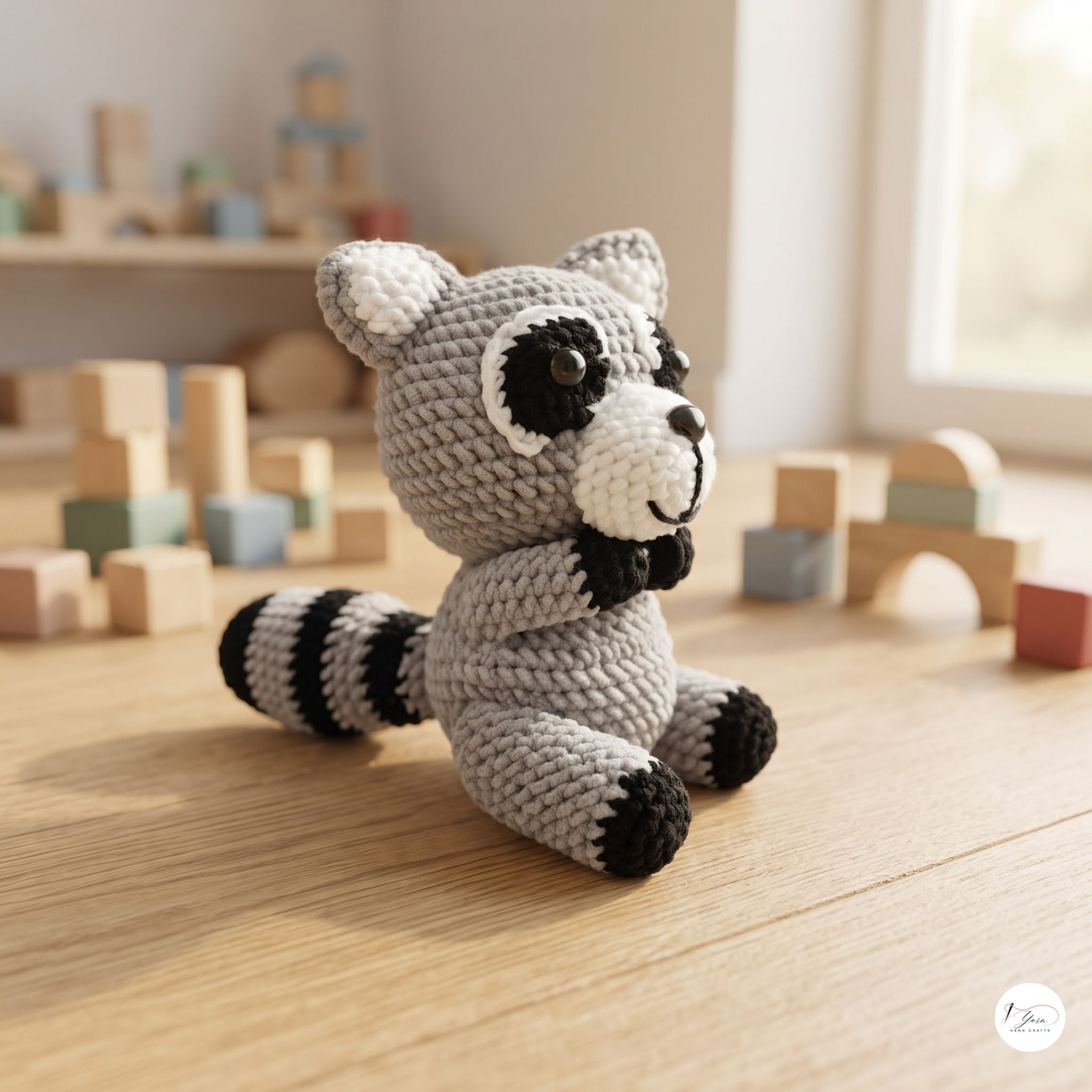 A crocheted raccoon toy sits on a light wood floor in a softly blurred children's room setting with wooden blocks in the background.