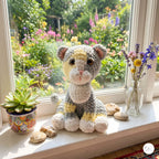 A crocheted stuffed animal cat with yellow, gray, and white coloring sits on a white windowsill. The background shows a lush, colorful garden visible through the window, with a small potted succulent on the left and a glass vase of flowers on the right.