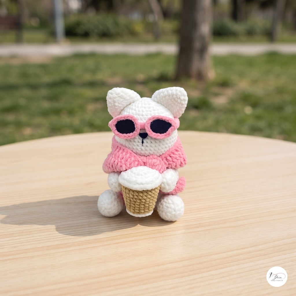 A crocheted white bunny wearing pink sunglasses and a pink scarf, sitting on a light wooden table outdoors with a blurred green park background.