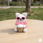 A crocheted white bunny wearing pink sunglasses and a pink scarf, sitting on a light wooden table outdoors with a blurred green park background.