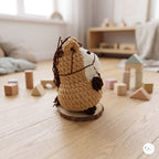 A crocheted plush horse toy sits on a round wooden coaster on a light wood floor in a softly lit nursery or playroom. The toy is tan with a white face, dark brown mane and hooves, and is surrounded by wooden blocks and toys in the background.
