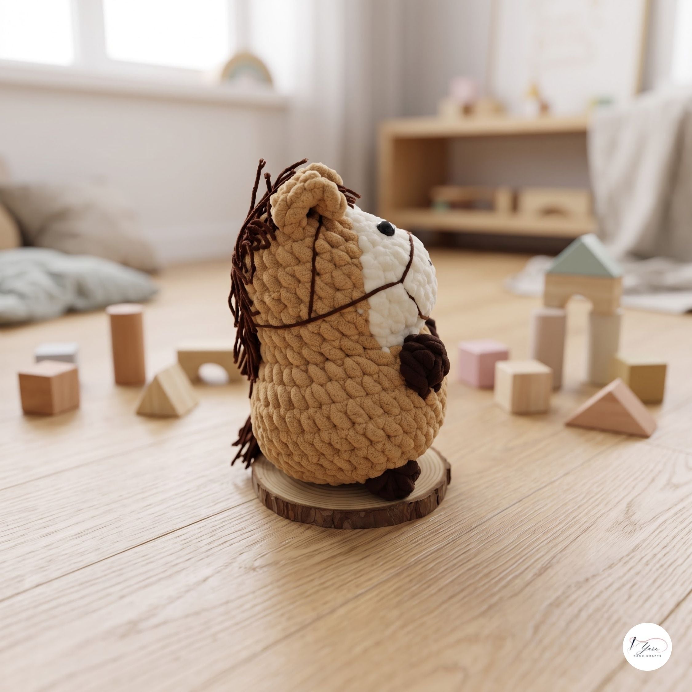 A crocheted plush horse toy sits on a round wooden coaster on a light wood floor in a softly lit nursery or playroom. The toy is tan with a white face, dark brown mane and hooves, and is surrounded by wooden blocks and toys in the background.