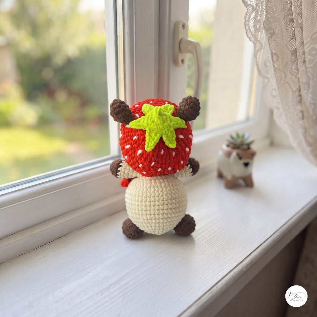 A crocheted strawberry gnome doll sits on a white window sill, facing away from the camera. The doll has a red and white strawberry hat with a green leafy top, brown ears and feet, and a cream-colored body.