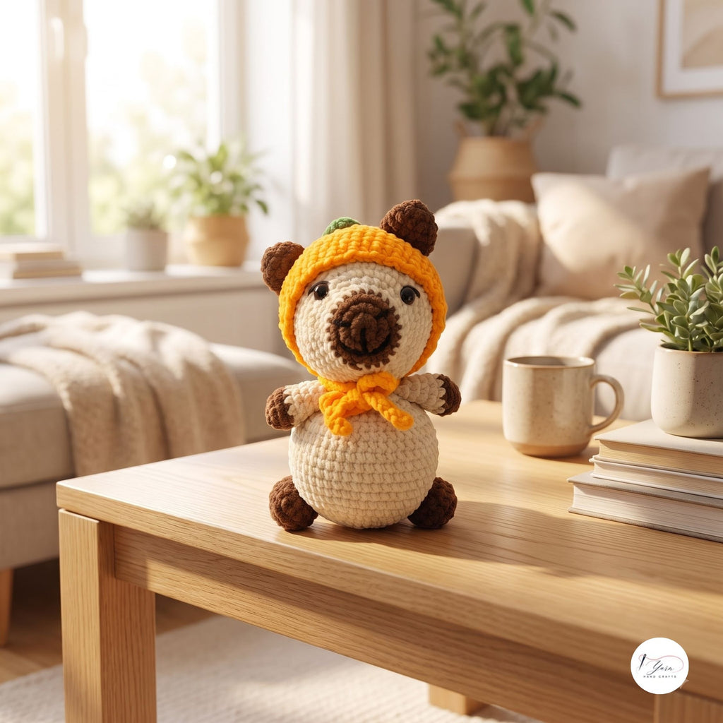 A crocheted plush bear toy wearing a bright yellow-orange hood and matching scarf, sitting on a light wood table in a cozy living room setting.