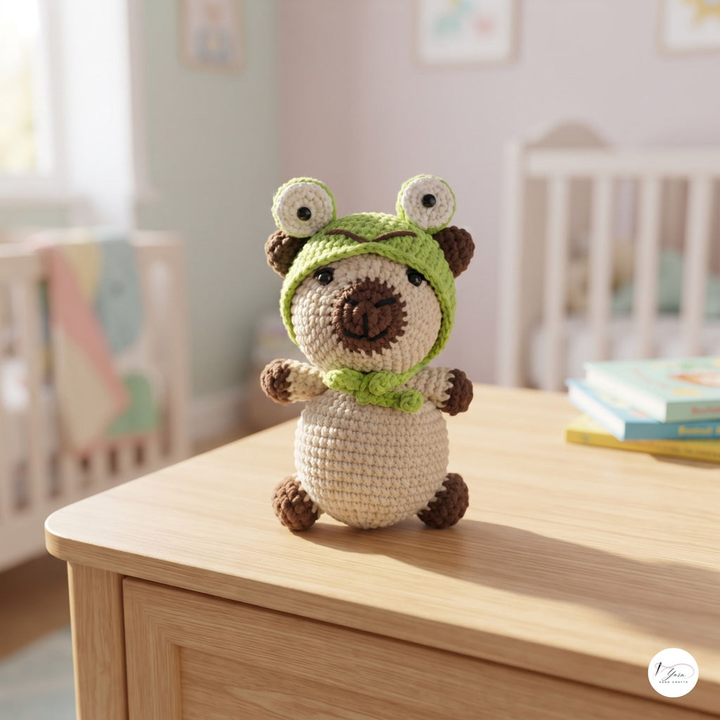 A crocheted amigurumi plush toy shaped like a bear wearing a green frog hood and matching green scarf, standing on a light wood table in a softly blurred nursery setting.