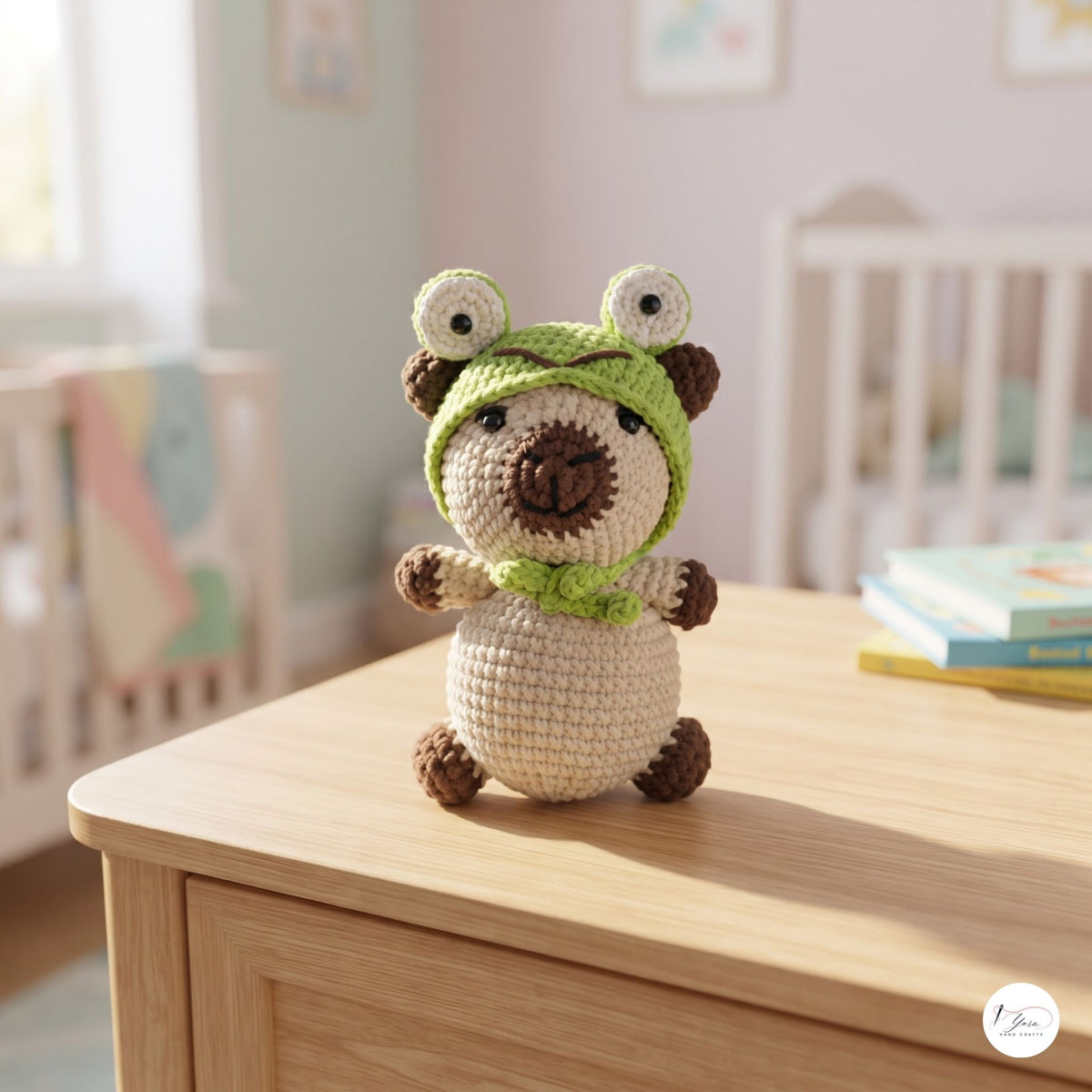 A crocheted amigurumi plush toy shaped like a bear wearing a green frog hood and matching green scarf, standing on a light wood table in a softly blurred nursery setting.
