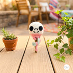 A crocheted amigurumi-style character ornament or figurine stands on a wooden deck outdoors, with potted plants and patio furniture blurred in the background. The character has a white head, black embroidered eyes and mouth, a pink stitched body, and a small mint-green accent.