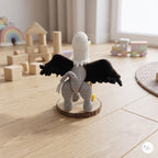 A handmade crocheted eagle figurine with outstretched dark brown wings, standing on a round wooden slice base. The figurine is photographed in a softly lit children's playroom with wooden blocks, a toy train, and a rainbow visible in the blurred background.