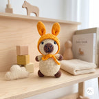 A crocheted amigurumi bunny doll with long orange-brown ears, a cream body, and a matching orange-brown bow sits on a light wood shelf styled with wooden blocks, a cloud patch, and a baby book.