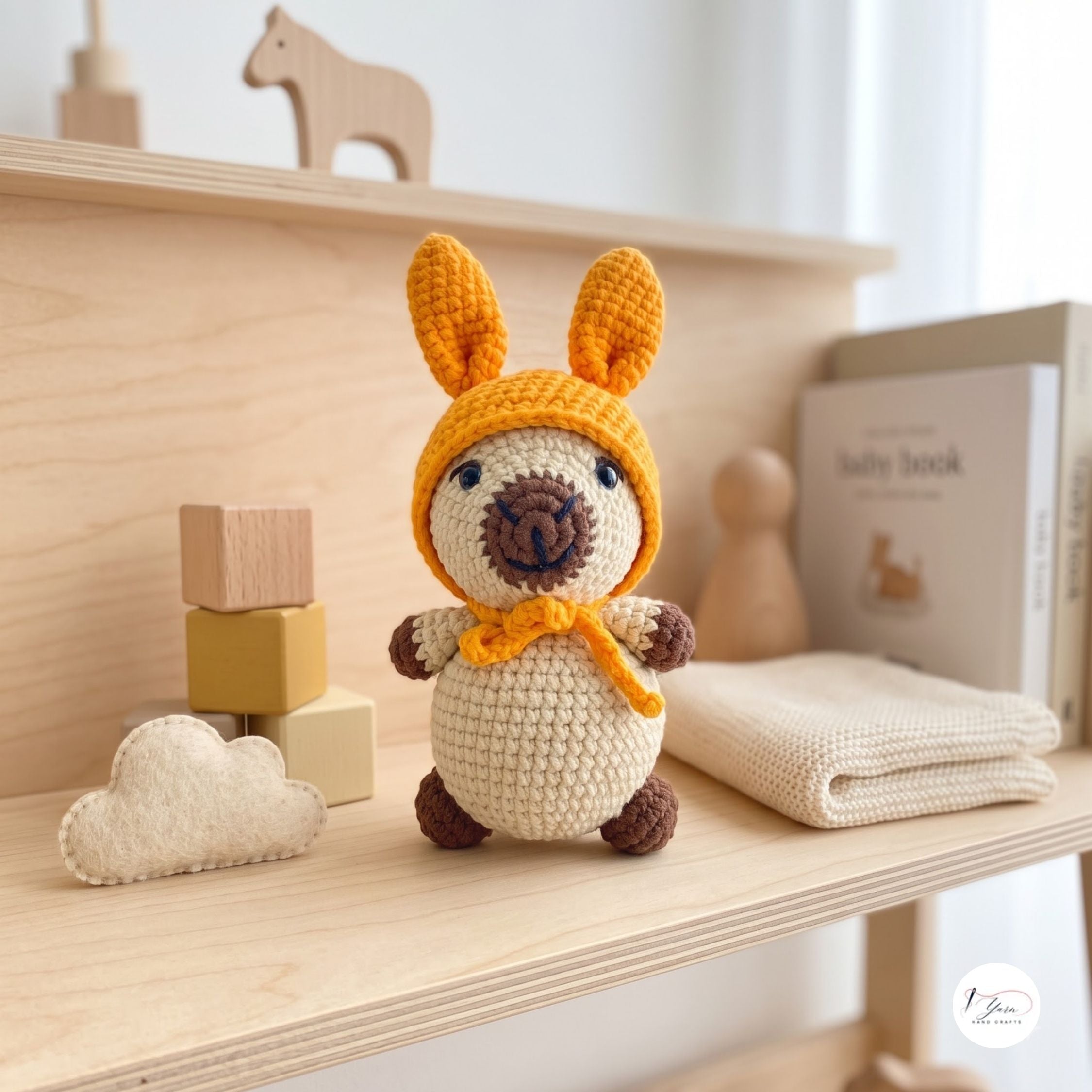 A crocheted amigurumi bunny doll with long orange-brown ears, a cream body, and a matching orange-brown bow sits on a light wood shelf styled with wooden blocks, a cloud patch, and a baby book.