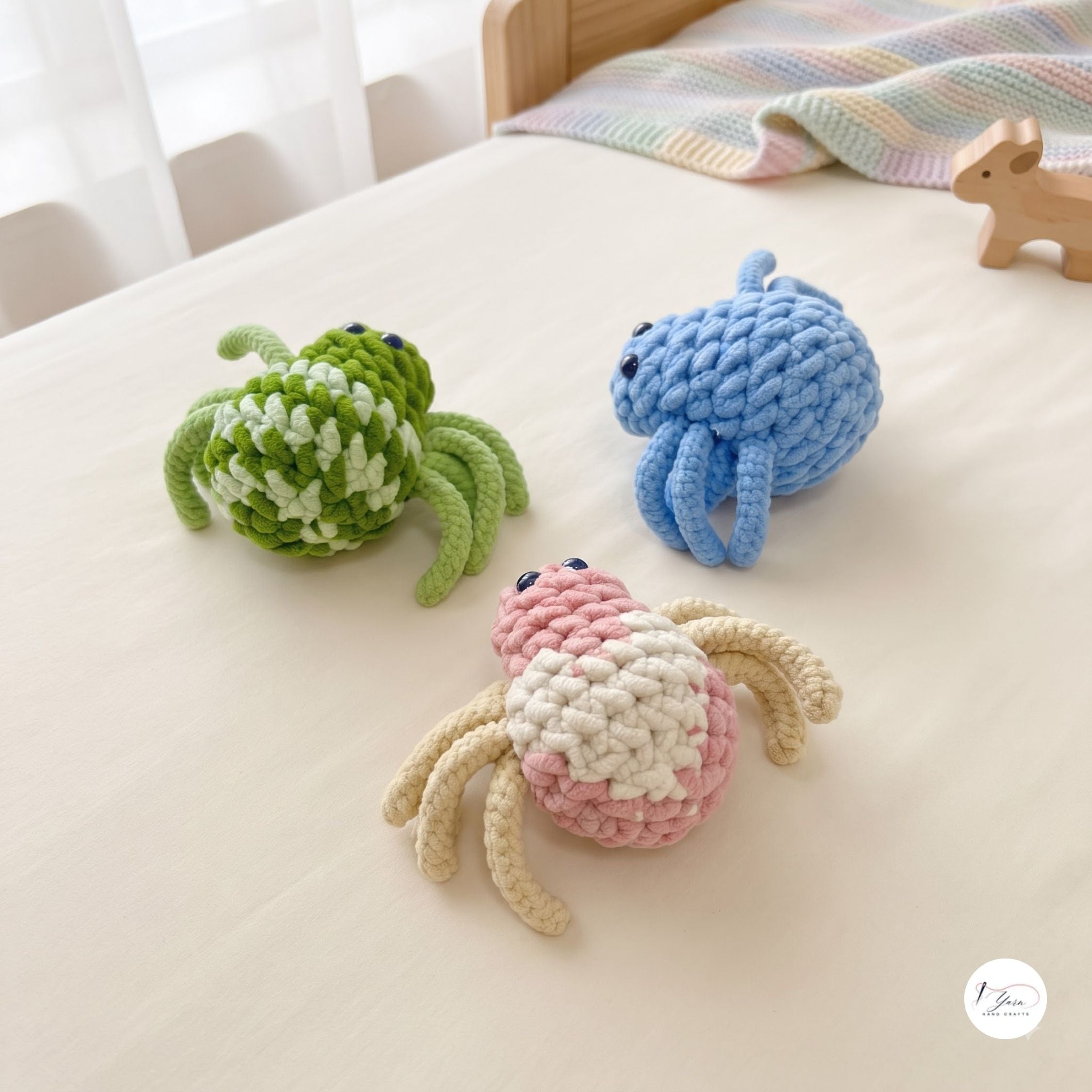A colorful trio of plush handmade crochet spider toys in green and cream, light blue, and pink and cream resting on a soft white bed sheet near a wooden bed frame with a pastel blanket and wooden toy.