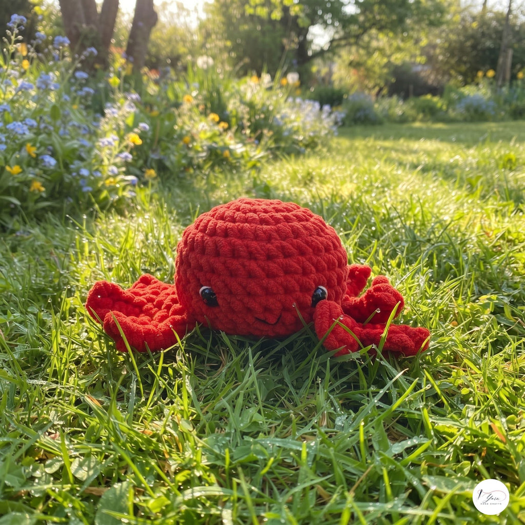 A red crocheted crab toy with small black bead eyes, lying on a green grassy lawn in a sunny garden.
