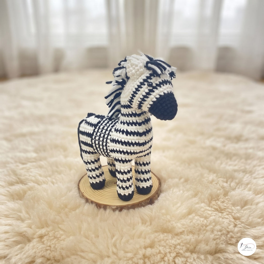 A crocheted zebra toy standing on a round wooden slice, placed on a fluffy cream rug with a softly blurred indoor background.
