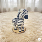 A crocheted zebra toy standing on a round wooden slice, placed on a fluffy cream rug with a softly blurred indoor background.