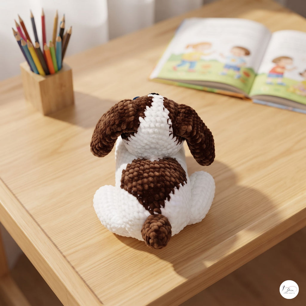 A crocheted dog toy with brown and white patches sits on a light wooden desk, with a pencil holder and an open book blurred in the background.