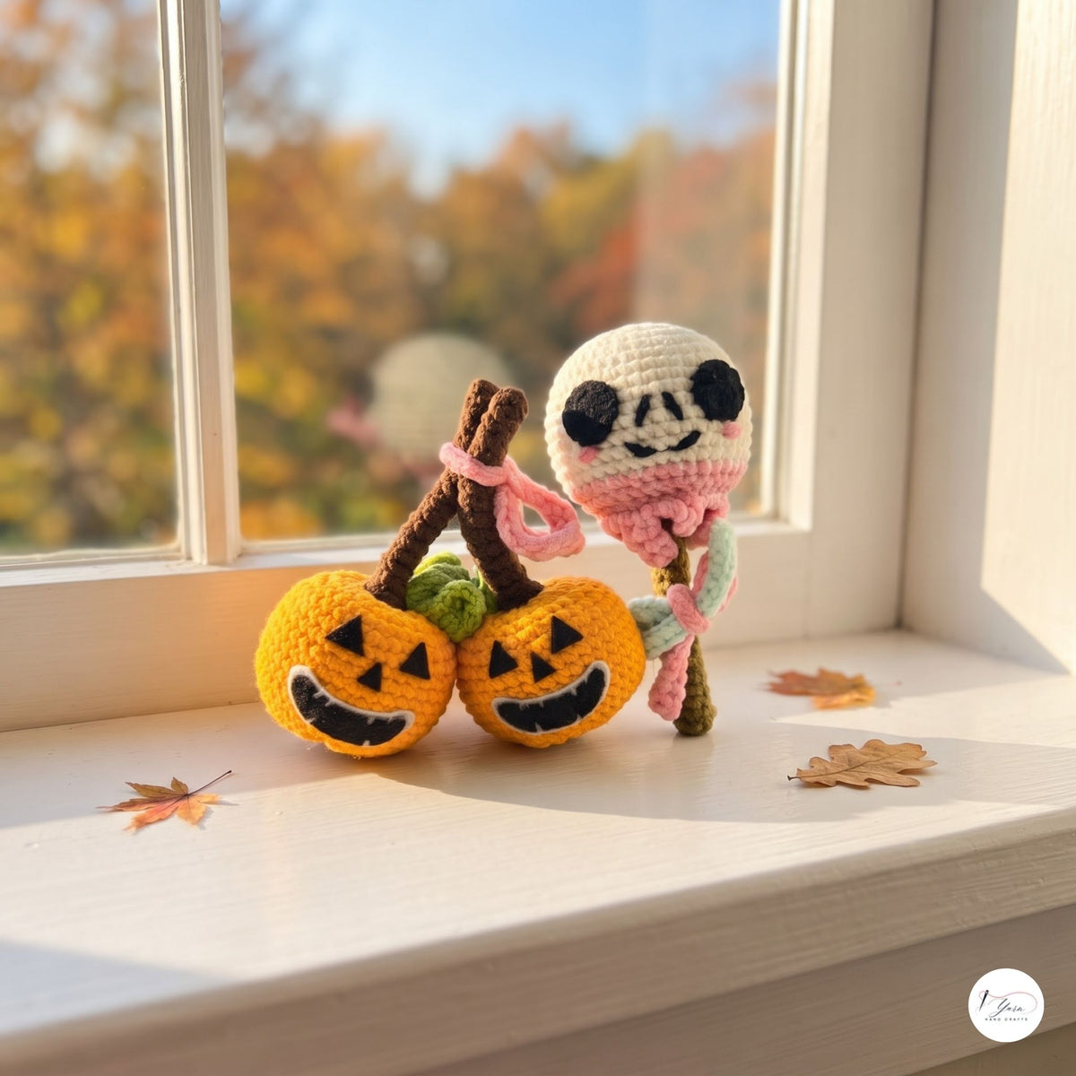 Handmade crocheted Halloween decoration set on a windowsill, featuring two orange jack-o'-lanterns with brown stems and a white skeleton figure with pink and green details, with autumn leaves scattered nearby.